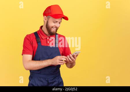 Portrait d'un messager souriant satisfait ou d'un artisan debout avec un smartphone entre les mains, prenant la commande en ligne, étant prêt à travailler. Studio d'intérieur isolé sur fond jaune. Banque D'Images