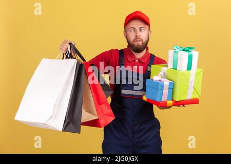 Drôle de courrier fatigué tenant beaucoup de boîtes présentes et sacs d'achats, posant avec les yeux croisés, ne sait pas comment gérer pour livrer la commande à temps. Studio d'intérieur isolé sur fond jaune Banque D'Images