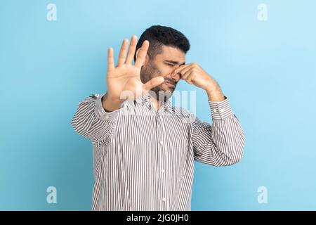 Un jeune homme d'affaires confus saisissant le nez avec les doigts et montrant un geste d'arrêt, se sentant une odeur dégoûtante désagréable, portant une chemise rayée. Studio d'intérieur isolé sur fond bleu. Banque D'Images