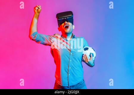Portrait d'un homme en chemise portant un casque et tenant le ballon de football, regardant l'équipe de football 3D, applaudisant, célébrant la victoire. Studio d'intérieur isolé sur fond de néon coloré. Banque D'Images