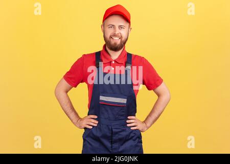 Portrait d'un travailleur confiant debout avec les mains sur les hanches, regardant l'appareil photo, étant prêt à prendre le contrôle et à terminer la commande, portant une combinaison et une casquette. Studio d'intérieur isolé sur fond jaune Banque D'Images