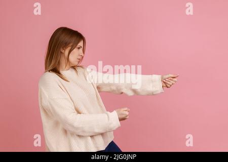 Portrait de femme blonde s'efforçant dur de tirer la corde invisible, tenant la lourde charge virtuelle avec l'expression de grand effort, portant chandail blanc. Studio d'intérieur isolé sur fond rose. Banque D'Images