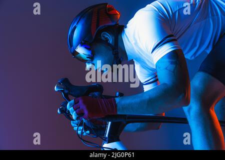 Gros plan portrait d'un cycliste professionnel en uniforme de sport, lunettes et casque sur fond bleu au néon. Concept de mode sport, course Banque D'Images