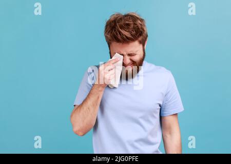 Portrait de malheureux frustré barbu homme socs et frotte les larmes avec mouchoir se sent désespéré, a insatisfait et expression de douleur. Studio d'intérieur isolé sur fond bleu. Banque D'Images