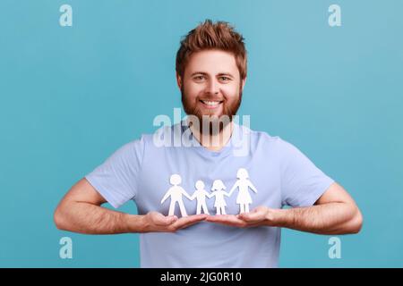 Portrait d'un beau barbu portant un T-shirt avec des personnes en chaîne de papier qui tiennent entre les mains, famille heureuse, relations, parentalité. Studio d'intérieur isolé sur fond bleu. Banque D'Images