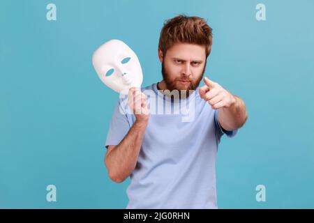 Portrait d'un jeune homme barbu adulte agressif et strict tenant le masque blanc à la main, pointant le doigt vers l'appareil photo avec une expression bossy sérieuse. Studio d'intérieur isolé sur fond bleu. Banque D'Images