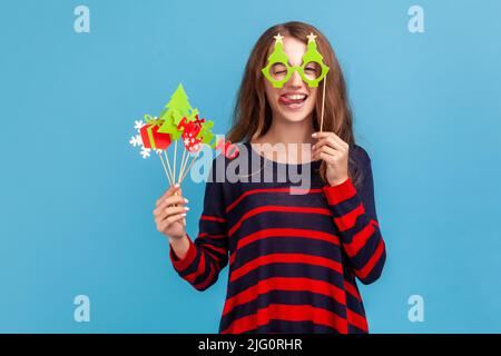 Drôle positive femme portant un chandail rayé style décontracté, tient les accessoires de fête de Noël, couvre les yeux avec des lunettes de papier, montre la langue dehors, humeur de fête. Studio d'intérieur isolé sur fond bleu Banque D'Images