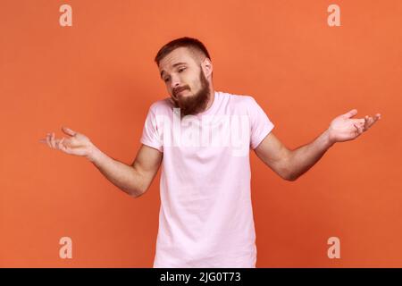 Ne sais pas, qui s'en soucie. Portrait d'un homme barbu debout sans geste d'idée, haussant les épaules en levant les mains, portant un T-shirt rose. Studio d'intérieur isolé sur fond orange. Banque D'Images