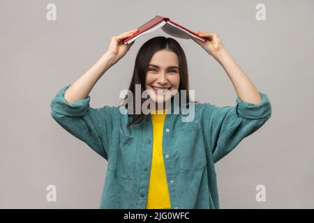 Drôle de femme positive tenant le livre ouvert sur la tête, étudiant faisant le rituel de se rappeler beaucoup de matériel d'étude, portant la veste de style décontracté. Prise de vue en studio isolée sur fond gris. Banque D'Images