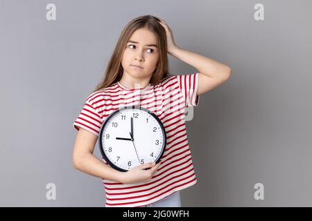 Portrait d'une petite fille confuse et incertaine portant un T-shirt rayé tenant une grande horloge murale entre les mains, regardant loin avec une expression perplexe. Prise de vue en studio isolée sur fond gris. Banque D'Images