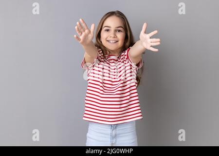 Entrez dans mes bras. Portrait d'une fillette souriante portant un T-shirt rayé qui dépasse les mains pour embrasser, offrant des hugs libres et accueillants. Prise de vue en studio isolée sur fond gris. Banque D'Images