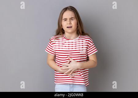 Portrait d'une petite fille portant un T-shirt rayé encombrant le ventre, sensation d'inconfort ou de douleur dans l'estomac, enfant malade souffrant de crampes de constipation. Prise de vue en studio isolée sur fond gris. Banque D'Images