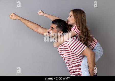 Gai Daddy piggyback fille, faisant voler la main super-héros, le geste avant de poing, petite fille à cheval les pères de retour, jouant le jeu actif ensemble. Prise de vue en studio isolée sur fond gris. Banque D'Images