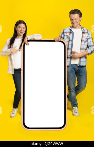 Pointant vers l'écran blanc homme caucasien et femme asiatique debout adossé sur un immense smartphone avec écran blanc, publicité d'application mobile isolée sur fond jaune. Positionnement du produit. Banque D'Images