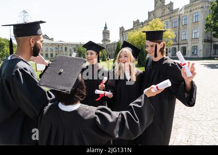 Baccalauréat multiethnique positif en casquettes et en robes détenant des diplômes dans le parc Banque D'Images