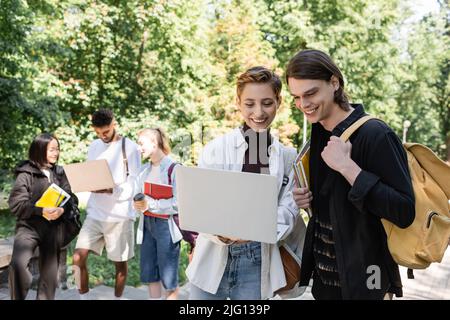 Des étudiants joyeux utilisant un ordinateur portable près d'amis multiculturels flous dans le parc Banque D'Images