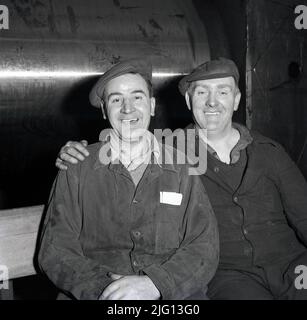 1950s, historique, deux ouvriers gallois de l'acier assis pour leur photo, souriant, dans leurs salopettes de travail et portant des casquettes en tissu, avec une feuille d'acier pressé sur un rouleau derrière eux. Photo prise à Abbey Works, Port Talbot, pays de Galles, Royaume-Uni, la maison de la compagnie d'acier du pays de Galles, à cette époque, le plus grand producteur d'acier et de platine en Europe. Banque D'Images