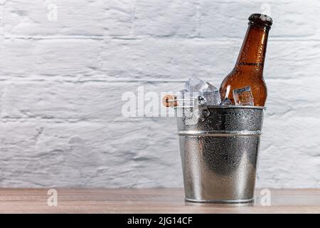 Seau en métal rempli de glace avec une bouteille de bière fraîche recouverte de gouttes de condensation sur une table en bois devant un mur blanc. Banque D'Images