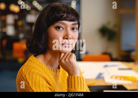 Portrait d'une jeune femme d'affaires biraciale confiante avec main sur le menton au bureau Banque D'Images
