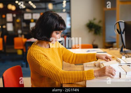 Vue latérale d'une jeune femme architecte biraciale examinant le modèle architectural dans un bureau créatif Banque D'Images