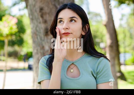 Jeune belle femme portant un t-shirt turquoise sur le parc de la ville, à l'extérieur touchant le menton réfléchi, pensant faire un choix important. En regardant vers le haut pendant que p Banque D'Images