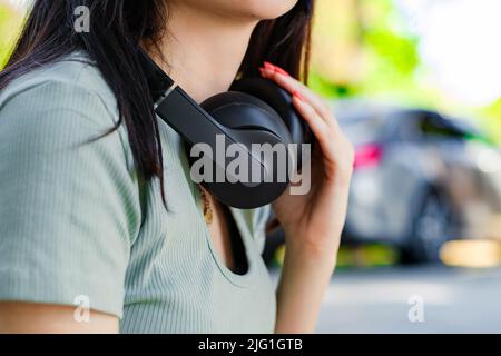 Brunette femme millénaire portant un t-shirt turquoise sur le parc de la ville, un casque d'extérieur autour de son cou et se touchant au casque. Banque D'Images