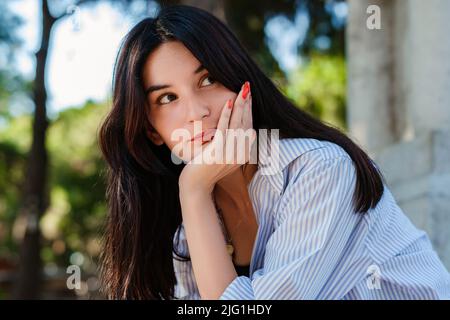 Adorable femme caucasienne portant une chemise rayée sur le parc de la ville, à l'extérieur touchant le menton réfléchi, pensant faire un choix important. Vue vers le haut Banque D'Images