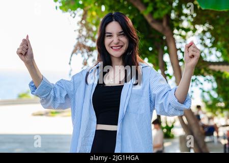 Jeune femme souriante confiante portant une chemise rayée debout sur le parc de la ville, plein air heureux et excités faisant le geste gagnant avec les bras relevés, sourire f Banque D'Images