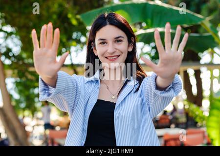 Gaie brune femme portant une chemise rayée bleue sur le parc de la ville, à l'extérieur montrant et pointant vers le haut avec les doigts numéro dix tout en souriant confiant et Banque D'Images