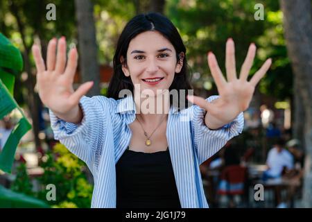 Gaie brune femme portant une chemise rayée bleue sur le parc de la ville, à l'extérieur montrant et pointant vers le haut avec les doigts numéro dix tout en souriant confiant et Banque D'Images