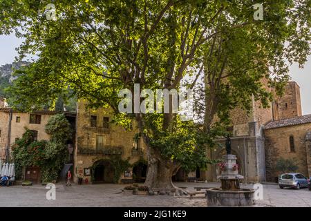 Des femmes se promènent sur la Place de la Liberté ombragée à Saint-Guilhem-le-Désert, Hérault, où un platane centenaire domine la fontaine de la place du village. Guilhem, cousin de Charlemagne, fonda l'abbaye de Gellone ici vers 804. Charlemagne lui offrit une relique de la Vraie Croix, encore portée en procession annuelle le 3 mai à travers le village. Le village se trouve sur la Via Tolosana, la route d'Arles du chemin de Saint-Jacques, est inscrit au registre des « Plus Beaux Villages de France » et fait partie du patrimoine mondial de l'UNESCO depuis 1998. Banque D'Images