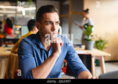 Jeune homme d'affaires caucasiens créatif et réfléchi, assis avec la main sur le menton au bureau Banque D'Images