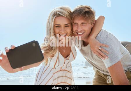 Un jeune couple aimant prend le selfie avec le téléphone tout en appréciant une journée à la plage souriante et endiabée. Romantique mari et femme montrant l'affection et Banque D'Images
