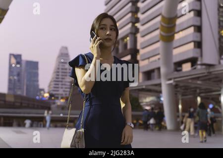 Jeune femme asiatique élégante et couronnée de succès dans des vêtements décontractés attendant quelqu'un et parlant au téléphone dans la rue de ville pendant la soirée Banque D'Images