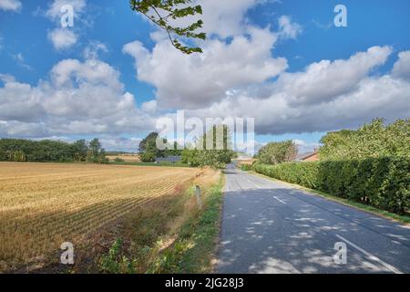 Paysage rural dynamique d'une route à travers la campagne par une journée ensoleillée. Champ sec de maïs ou de blé après récolte dans un petit village danois contre a Banque D'Images