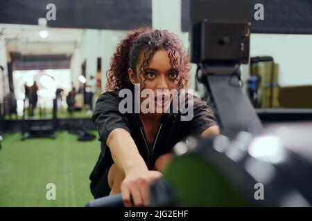 Femme afro-américaine utilisant une machine à ramer pendant l'entraînement en cross-fit. Une athlète féminine s'entraînant intensément dans la salle de sport. Photo de haute qualité Banque D'Images