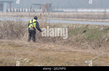 Lubaczow, Pologne - 9 mars 2022: Nettoyage des fossés de drainage en hiver. Faucher de l'herbe avec une débroussailleuse. Travailleur municipal au travail. Banque D'Images