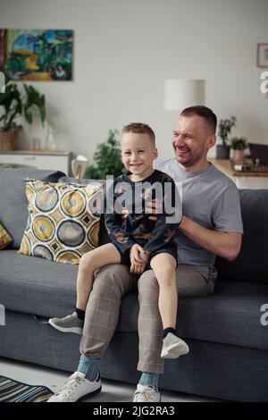 Joyeux petit garçon assis à genoux de son père et jouant avec lui sur le canapé dans le salon Banque D'Images