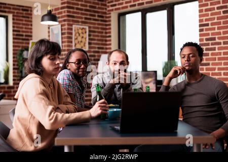Groupe multiracial de personnes assis à la maison tout en regardant des films de suspense sur ordinateur portable. Des personnes diverses à table dans le salon regardent des contenus vidéo en ligne ensemble tout en prenant des en-cas et des boissons. Banque D'Images