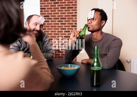 Homme afro-américain jouant au jeu de devinettes avec des cartes tout en sirotant de la bière avec des amis à la maison. Heureux souriant amis multiraciaux assis à table dans le salon tout en appréciant les activités de loisirs. Banque D'Images