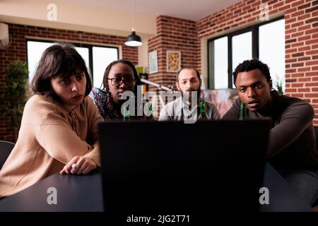 Diverses personnes assises à table dans le salon tout en regardant des films dramatiques ensemble sur un ordinateur portable. Groupe multiracial d'amis assis à la maison tout en appréciant des films de suspense. Banque D'Images