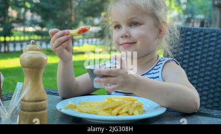 Une petite fille mange des frites. Gros plan de blonde fille prend des chips de pomme de terre avec ses mains et les essaie assis dans la rue café sur le parc. Banque D'Images