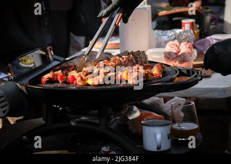 Restaurant de restauration rapide extérieur dans le parc. Rôti de viande de kebab et légumes grillés. Le chef en gants noirs rôde la viande sur le gril et la retourne à l'aide de moi Banque D'Images