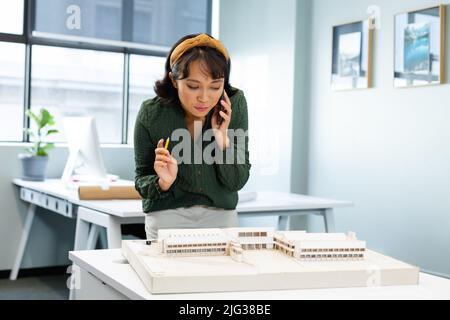 Jeune architecte asiatique parlant sur téléphone mobile tout en examinant le modèle architectural dans le bureau Banque D'Images