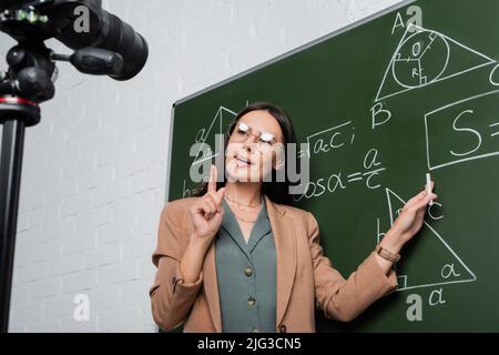 Professeur de lunettes expliquant les formules mathématiques sur tableau de surveillance près d'un appareil photo numérique en classe Banque D'Images