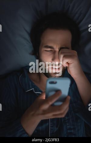 Vue de dessus de l'homme épuisé dans le pyjama avec un smartphone sur le lit la nuit Banque D'Images
