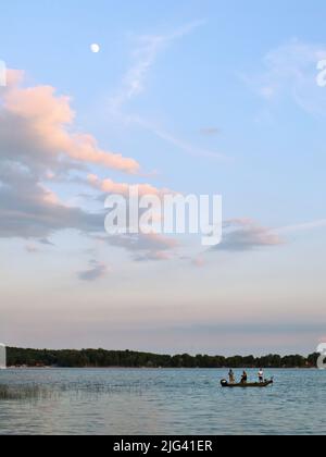 La lune et les nuages spectaculaires bordés d'orange par le soleil couchant offrent une toile de fond paisible pour trois hommes qui pêchent sur un lac. Banque D'Images