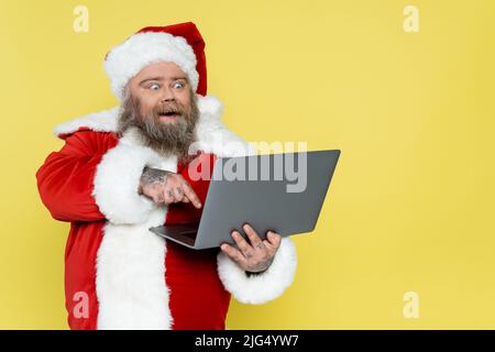 le père noël en surpoids stupéfié pointe vers un ordinateur portable isolé sur du jaune Banque D'Images