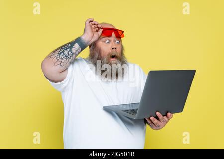 surpris homme de taille plus tenant des lunettes de soleil rouges tout en regardant l'ordinateur portable isolé sur jaune Banque D'Images