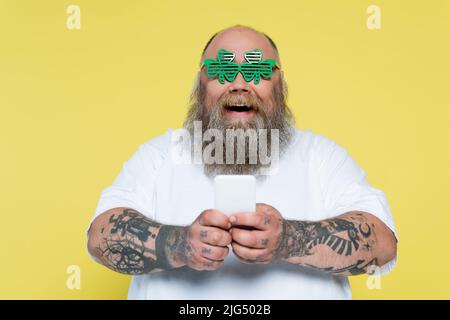 un homme de grande taille dans des lunettes en forme de trèfle bavardant sur un smartphone isolé sur le jaune Banque D'Images